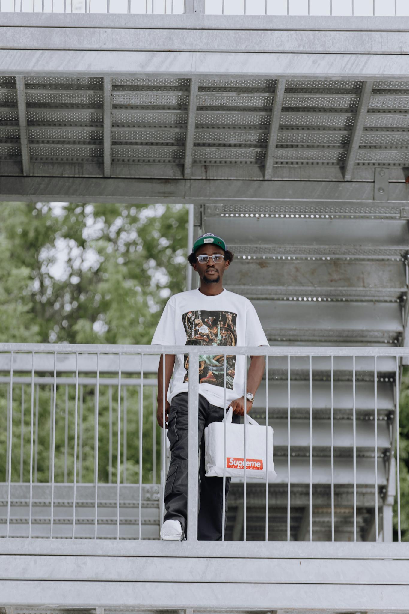 A young man in casual wear stands on a metal staircase holding a white bag.