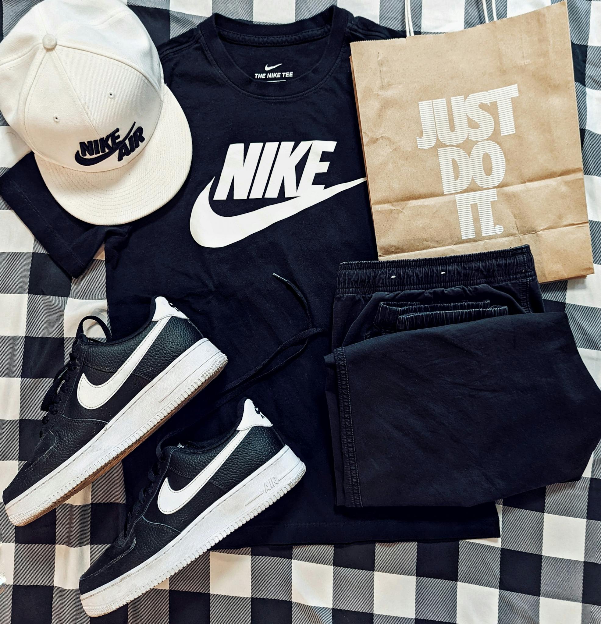 Flat lay of Nike apparel including t-shirt, cap, sneakers, and paper bag on checkered surface.