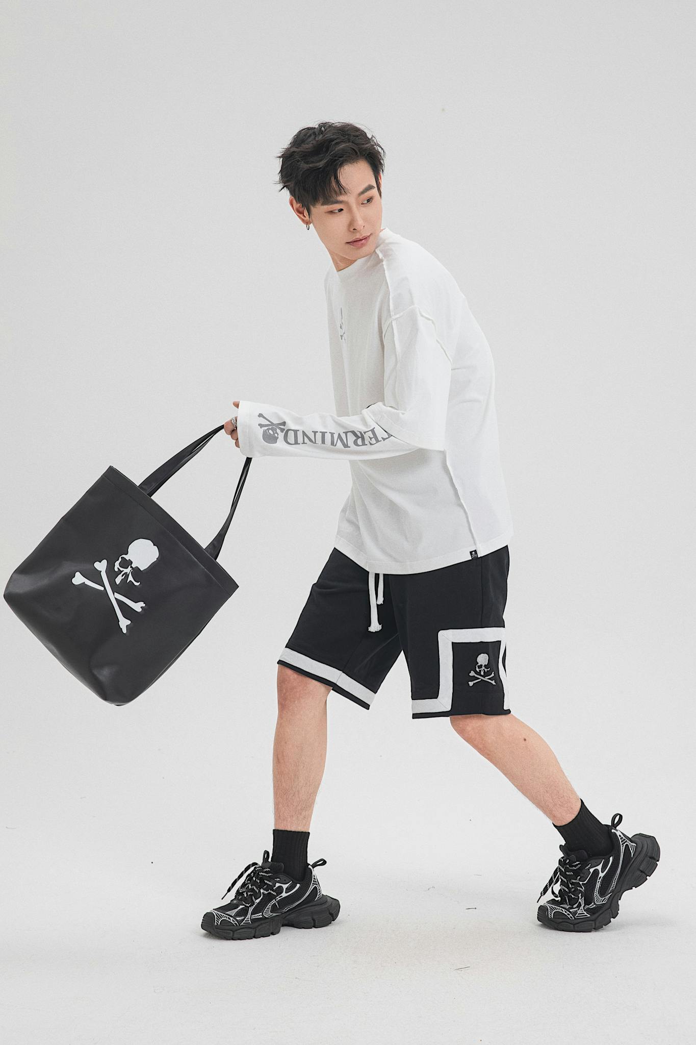 Male model posing in stylish sportswear with skull design tote bag in studio.